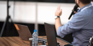 Tisch mit Laptops und Mann der einen Vortrag hält