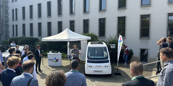 A small white autonomous shuttle is parked in a courtyard, about 30 people are standing in front of it and Jonathan Behm points to the cab at that moment