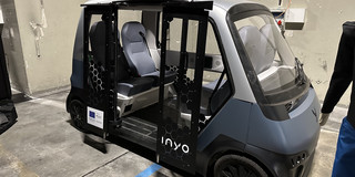 INYO Cab Zu sehen ist das INYO Cab mit geöffneten Türen. Es ist ein Shuttle von der Größe eines Golfkarts mit seitlichem Einstieg und vier sich gegenüberstehenden Stühlen.