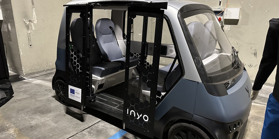 Zu sehen ist das INYO Cab mit geöffneten Türen. Es ist ein Shuttle von der Größe eines Golfkarts mit seitlichem Einstieg und vier sich gegenüberstehenden Stühlen.